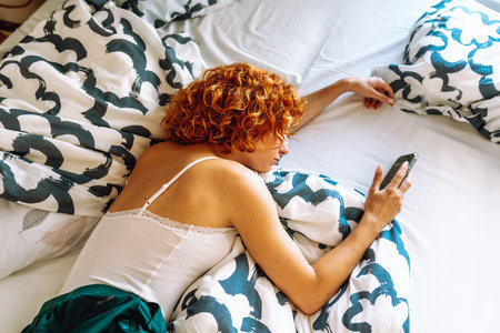 portrait of red-haired young woman lying on bed using mobile phoneの写真素材