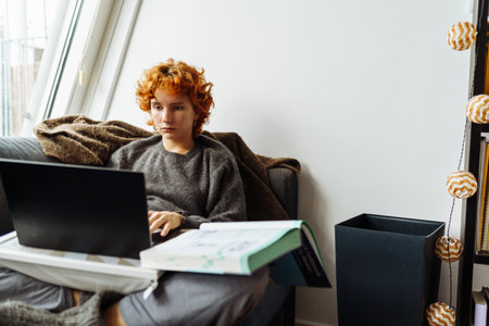 young red-haired woman sitting on soft sofa in warm sweater, using tablet, working, studying, e-learningの写真素材