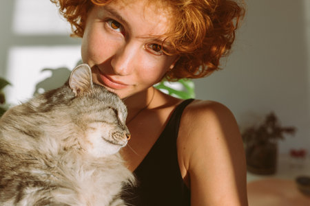 portrait red-haired curly young woman with pet gray fluffy cat in arms, lit by bright sun