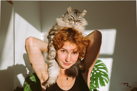 portrait red-haired curly young woman with pet gray fluffy cat in arms, lit by bright sunの写真素材