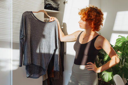 portrait of young red-haired woman choosing clothes in closet at homeの写真素材