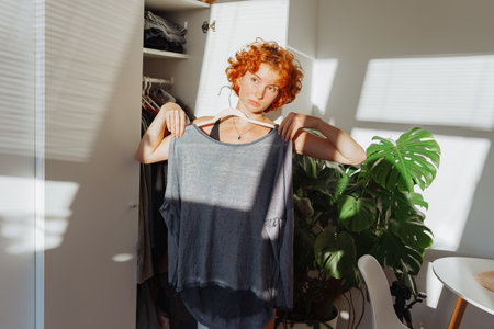 portrait of young red-haired woman choosing clothes in closet at home