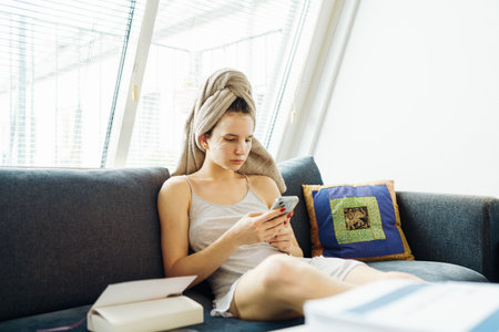 woman with towel on head using smartphone sitting on comfortable sofaの写真素材