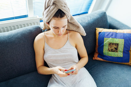 woman with towel on head using smartphone sitting on comfortable sofaの写真素材