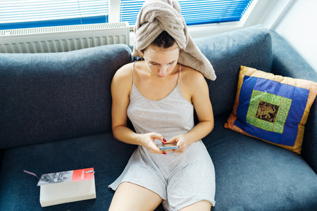 woman with towel on head using smartphone sitting on comfortable sofaの写真素材