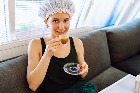 young woman with shower cap on head, sitting on soft sofa, enjoying cup of morning coffee with milkの写真素材