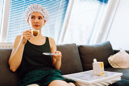 young woman with shower cap on head, sitting on soft sofa, enjoying cup of morning coffee with milkの写真素材