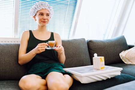 young woman with shower cap on head, sitting on soft sofa, enjoying cup of morning coffee with milkの写真素材