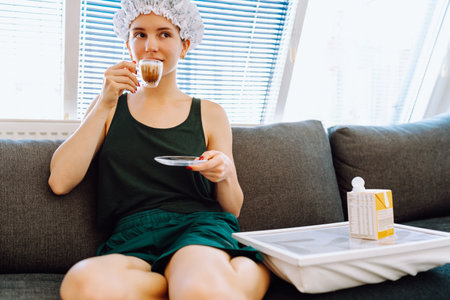 young woman with shower cap on head, sitting on soft sofa, enjoying cup of morning coffee with milkの写真素材
