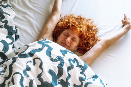 young attractive redhead curly woman waking up with pleasure stretching lying in bedの写真素材