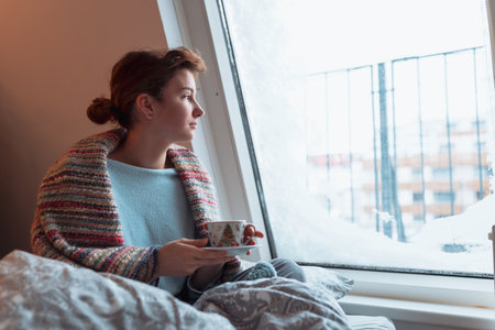 young woman warms up with hot tea in winter weather at homeの写真素材