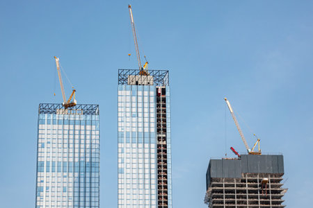Moscow, Russia - April 13, 2021: Skyscraper construction. Construction cranes on the roofs of three high-rise buildings under construction against the blue sky.のeditorial素材