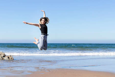 pretty young woman jumping for joy on the beach by the sea on a sunny blue day, wearing jeans