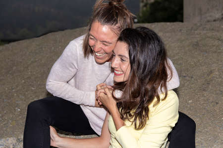 young lesbian couple laughing embracing sitting on a wall