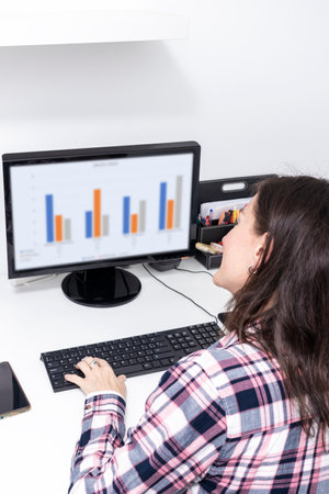 Close-up rear view of young business woman working in office interior on pc on desk, typing, looking at screen with diagramsの写真素材