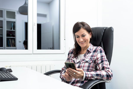 Mature woman using laptop whilst reading smartphone text at homeの写真素材