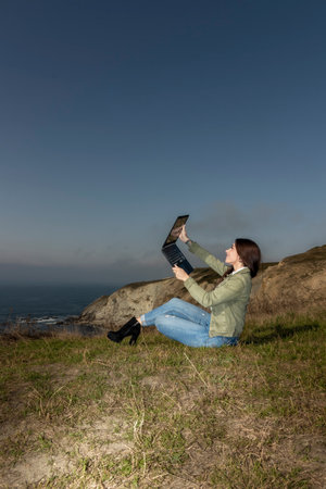Smiling Woman Embraces Nature While Engaging in a Laptop Video Call by the Seaの素材