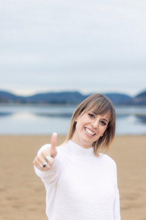 Radiant Beauty on the Beach: Smiling Woman in a White Sweater Giving a Thumbs Upの写真素材