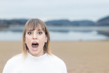 Captivating Beachside Surprise: Beautiful Woman in White Sweater Expresses Astonishment by the Seaの写真素材