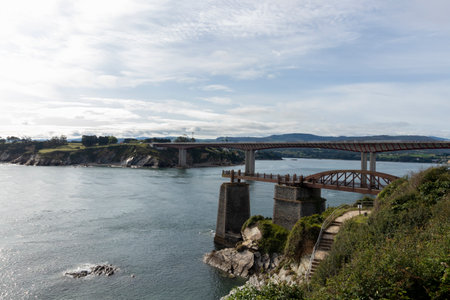 Serene View of a Curved Bridge Over a Calm River Under a Cloudy Skyの写真素材