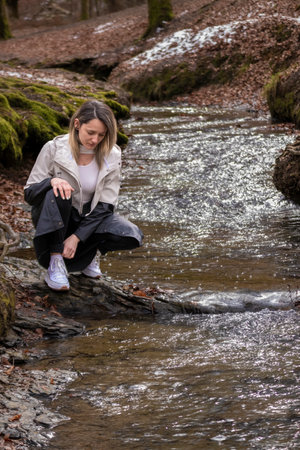 woman in white jacket and black pants is crouching on a rock in a forest streamの写真素材