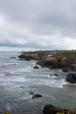 rocky coastline with a cloudy sky, rough waters, and a small lighthouse in the backgroundの写真素材