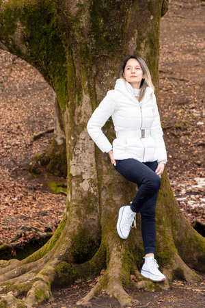 woman sitting on a moss-covered tree trunk in a forest, wearing a white jacket and black pantsの写真素材