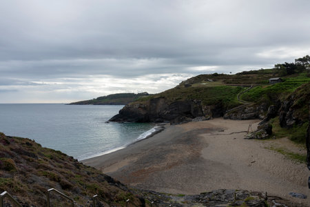 beach cove with a rocky cliff, a small building on top, and a cloudy sky overheadの写真素材