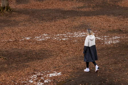 in a long coat and white sneakers walking in a park with fallen leaves and patches of snow on the groundの写真素材