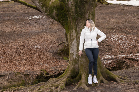 leaning against a tree in a snowy park, wearing a white jacket, blue jeans, and white sneakersの写真素材