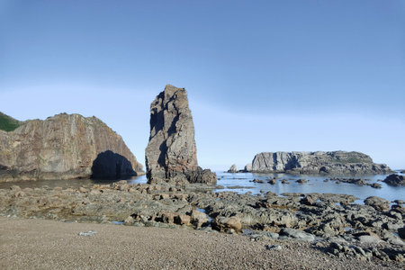 rocky beach with a tall, dark basalt formation in the center, surrounded by smaller rocks under a clear blue skyの写真素材