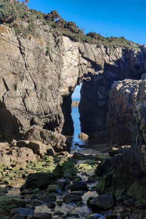 rocky beach features a tall, jagged arch formation, with the deep blue ocean visible through the arch under a clear skyの写真素材