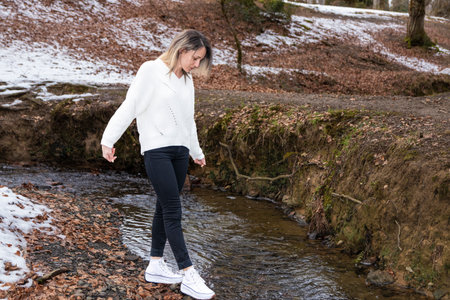 woman in a white sweater is crossing a small stream in a forest, stepping on rocks, with snow on the groundの写真素材