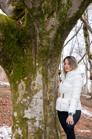 woman in a white jacket stands next to a moss-covered tree in a forestの写真素材