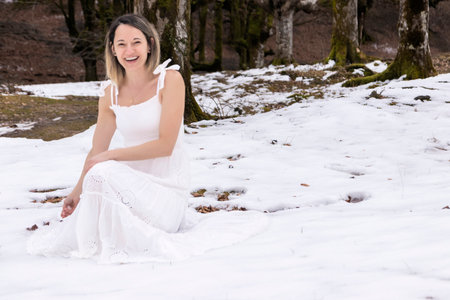 woman in a white, lace-patterned dress sits on a rock in a snowy, leafless forestの写真素材