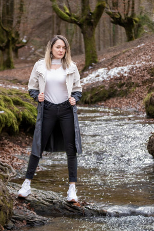 woman in a beige coat standing on a rock in a stream in a snowy forestの写真素材