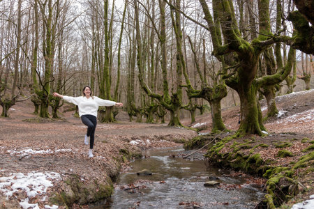 woman in a white sweater and black pants is joyfully jumping over a small stream in a forest with patches of snowの写真素材