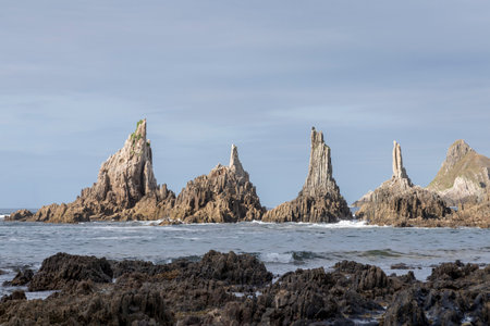 rocky coastline with three prominent, jagged rock formations in the ocean, under a clear sky. The foreground shows dark, barnacle-covered rocksの写真素材