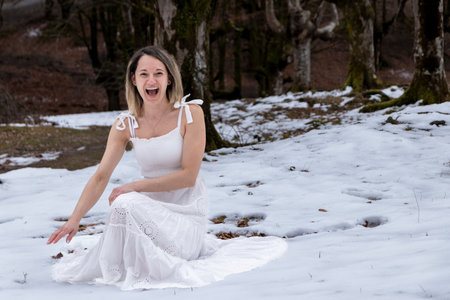 woman in a white, flowing dress with a lace pattern is sitting on a rock in a snowy forest, creating a peaceful and serene moodの写真素材