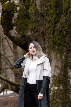 woman in a white shirt and gray jacket standing in front of a mossy tree in a snowy forestの写真素材