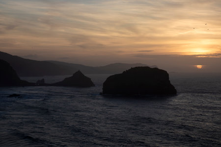 sunset over the ocean with a large rock formation silhouetted against the vibrant skyの写真素材