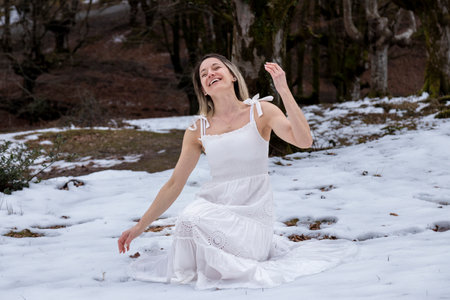 woman in a white, flowing dress sitting on a rock in a snowy forest with bare treesの写真素材