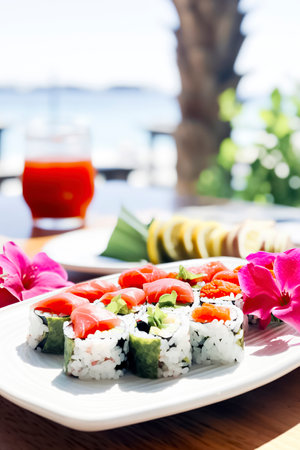 a plate of sushi with various rolls, garnished with flowers and lemon, accompanied by a red drink, against an ocean backdropの素材