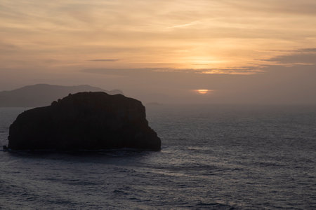 silhouette of a rocky island in the ocean, bathed in the warm hues of a setting sunの写真素材