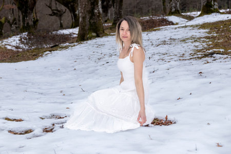 woman in a white lace dress sits serenely on a snow-covered rock in a peaceful, snowy forestの写真素材