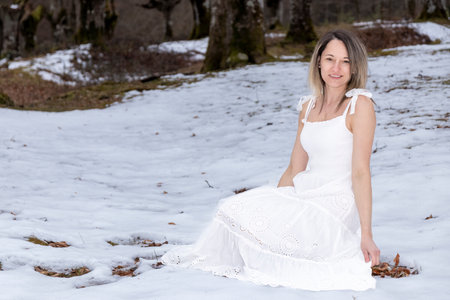 person in a long, flowing white dress sitting on a rock in a snowy forest with trees in the backgroundの写真素材