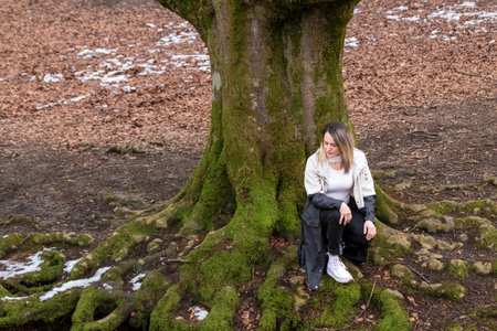 woman sitting on moss-covered tree roots in a forest, wearing a white shirt, black pants, and white shoesの写真素材
