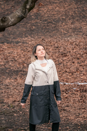 woman in a beige coat standing in a park with a tree branch and fallen leaves in the backgroundの写真素材