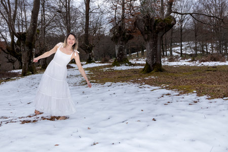 woman in a white dress dancing in a snowy forest, barefoot, with an overcast sky overheadの写真素材