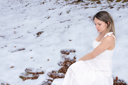 woman in a white dress sitting on a rock in a snowy forest, barefoot, with an overcast sky overheadの写真素材